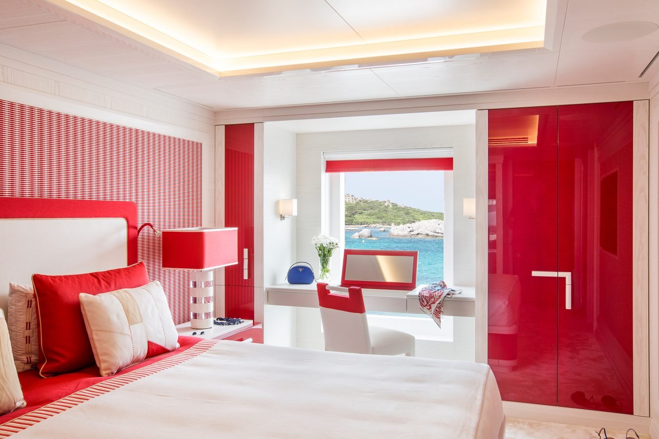 Modern bedroom with red and white decor, a large bed, desk, and window with ocean and greenery view outside.