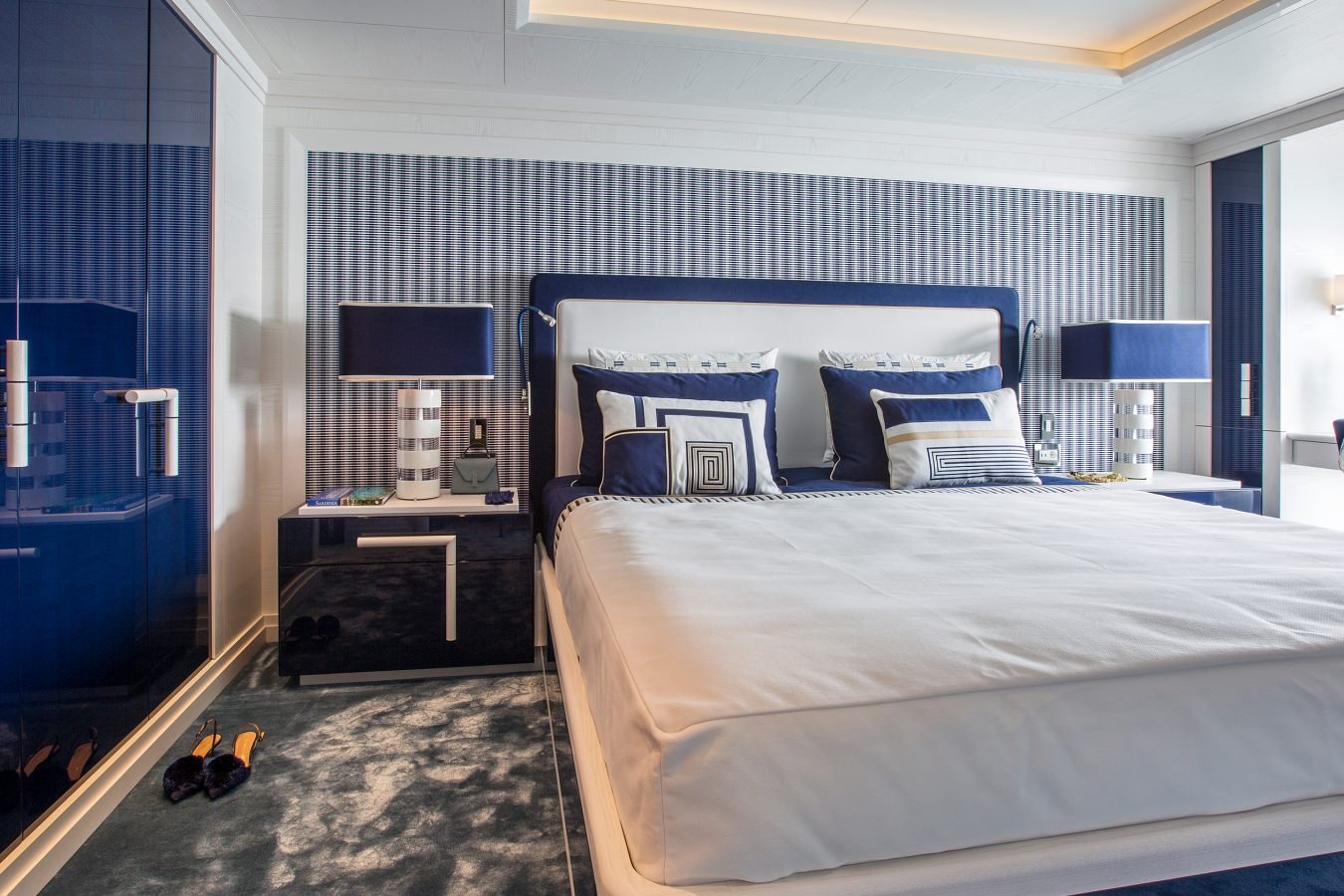 Modern bedroom with blue and white decor, large bed, matching nightstands, lamps, and a flower on the windowsill.