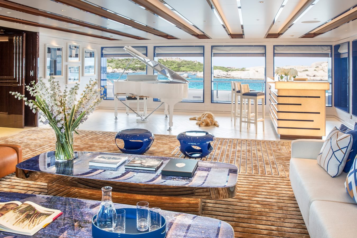 Spacious yacht lounge with modern decor, a white grand piano, sea views, blue accents, and a dog lying by the windows.