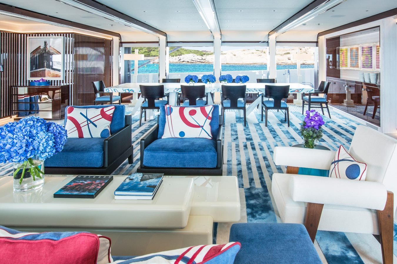 Modern yacht lounge with blue and white decor, abstract pillows, floral arrangements, and large windows with ocean view.