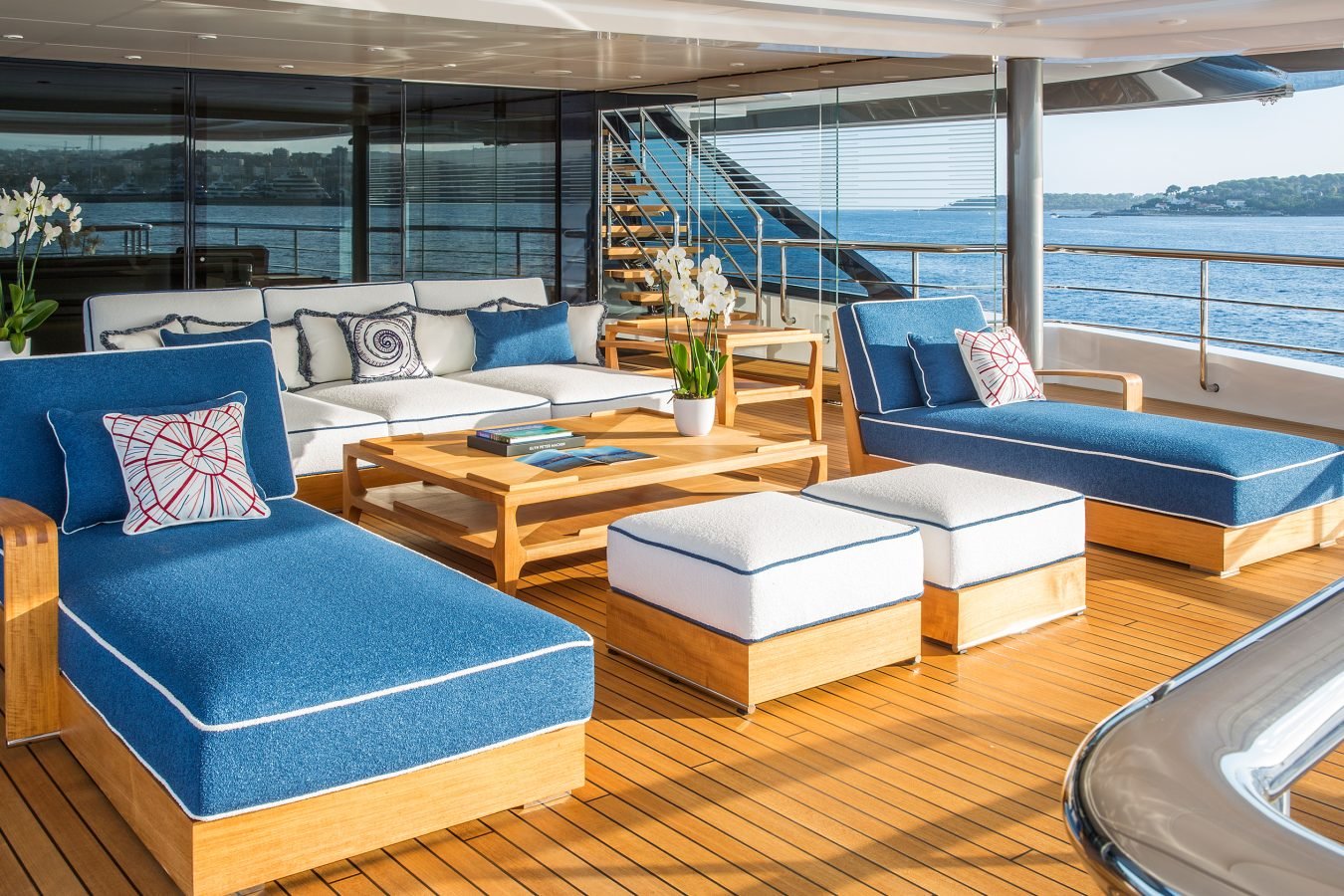 Elegant yacht deck with blue cushioned lounge chairs, ottomans, a sofa, tables, and scenic water view in the background.