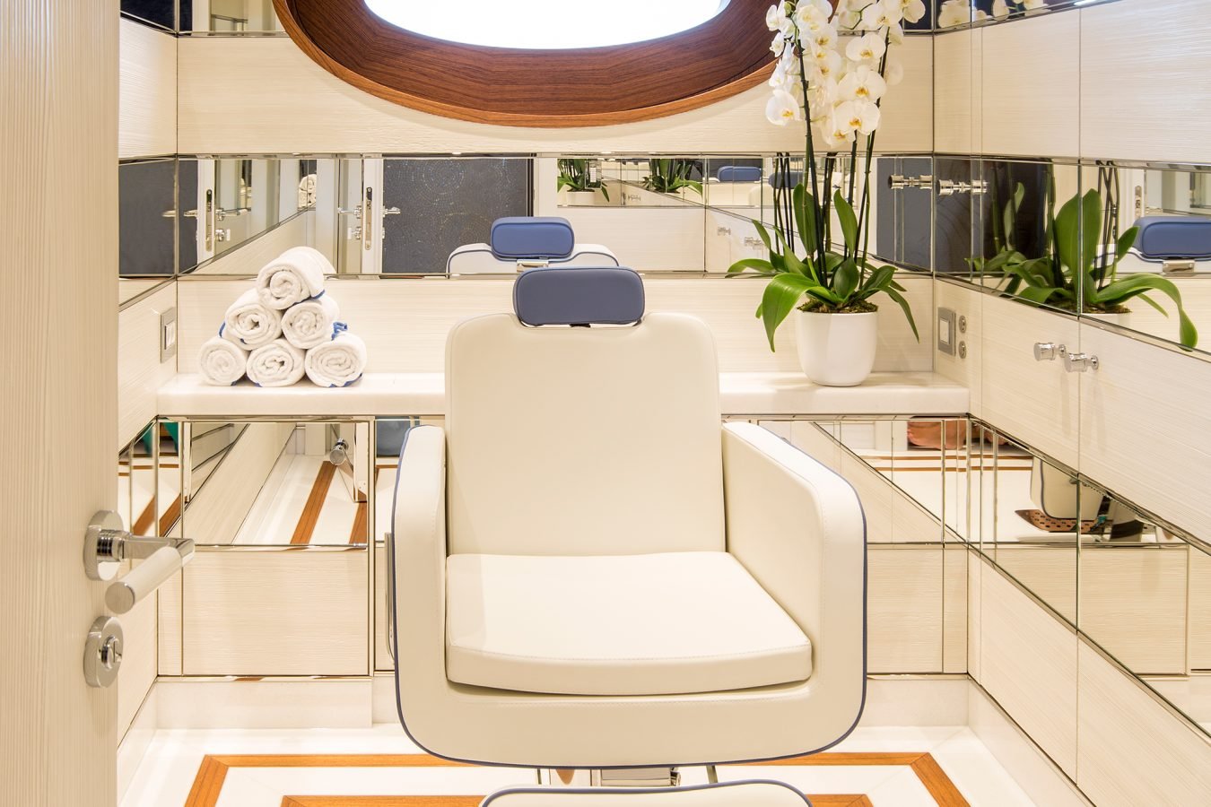 A modern, white salon chair faces mirrors in a bright, luxurious room with rolled towels and orchids on a counter.