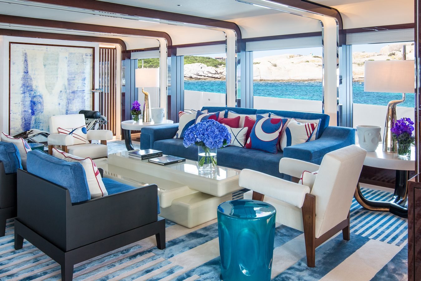 Modern yacht lounge with blue sofa, white armchairs, nautical pillows, fresh flowers, large windows, and ocean view.