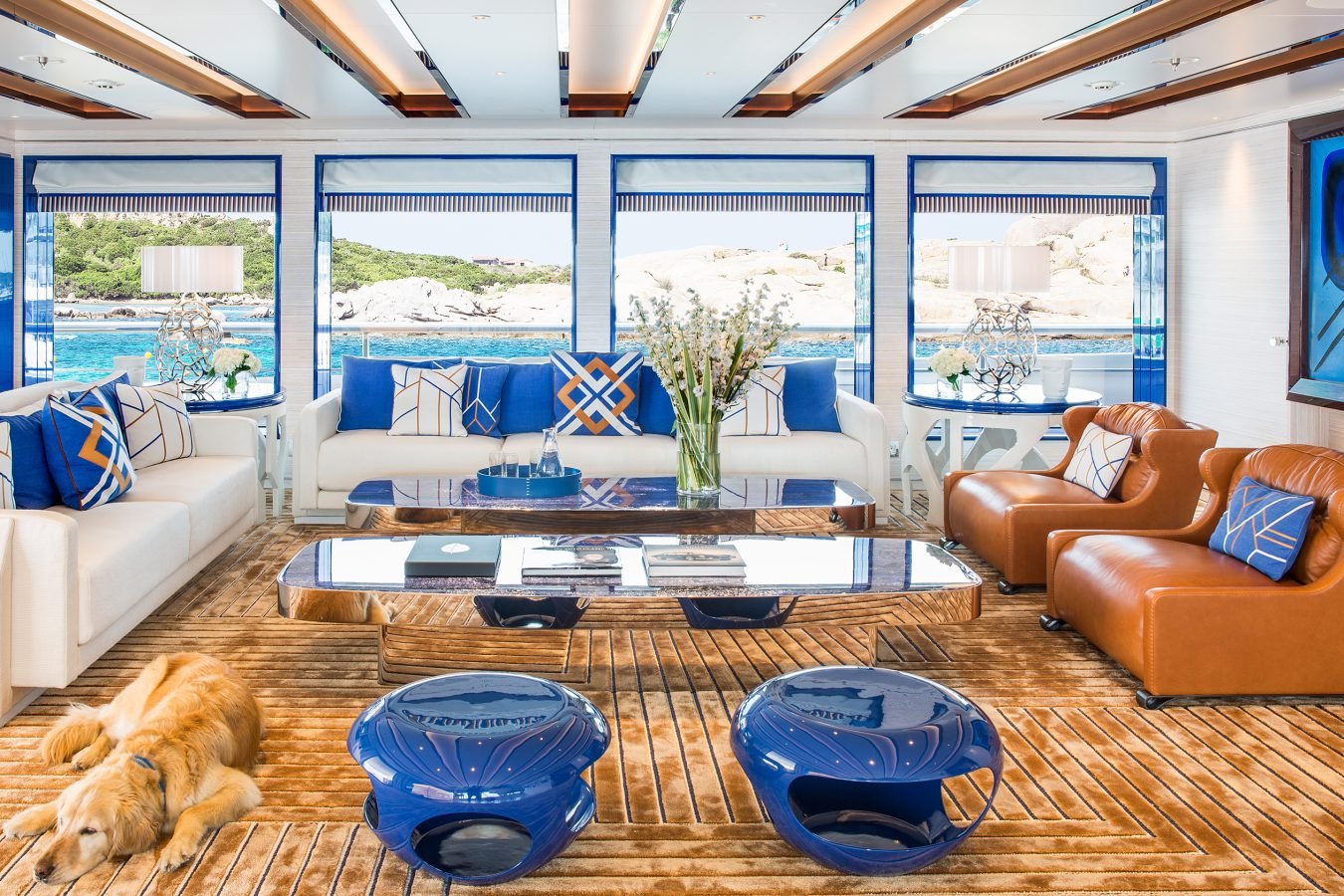 Modern yacht lounge with blue and white decor, tan chairs, glass table, sea view, and a dog lying on the wooden floor.