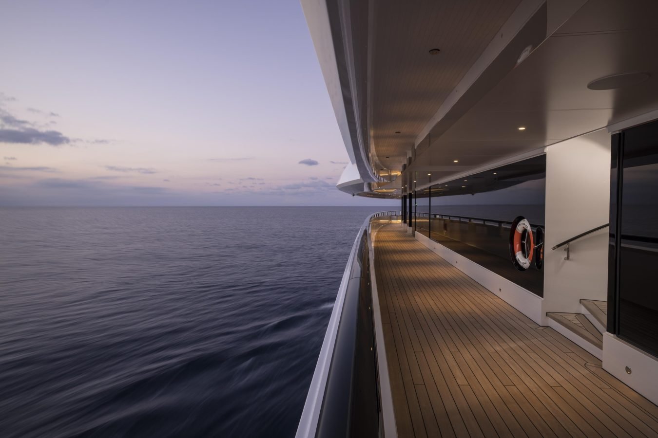 Modern yacht deck at sunset, with calm ocean, reflections on glass, and warm deck lights along the railings.