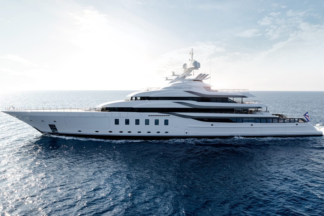 Large white luxury yacht cruising on calm blue ocean water under a clear sky with sunlight in the background.