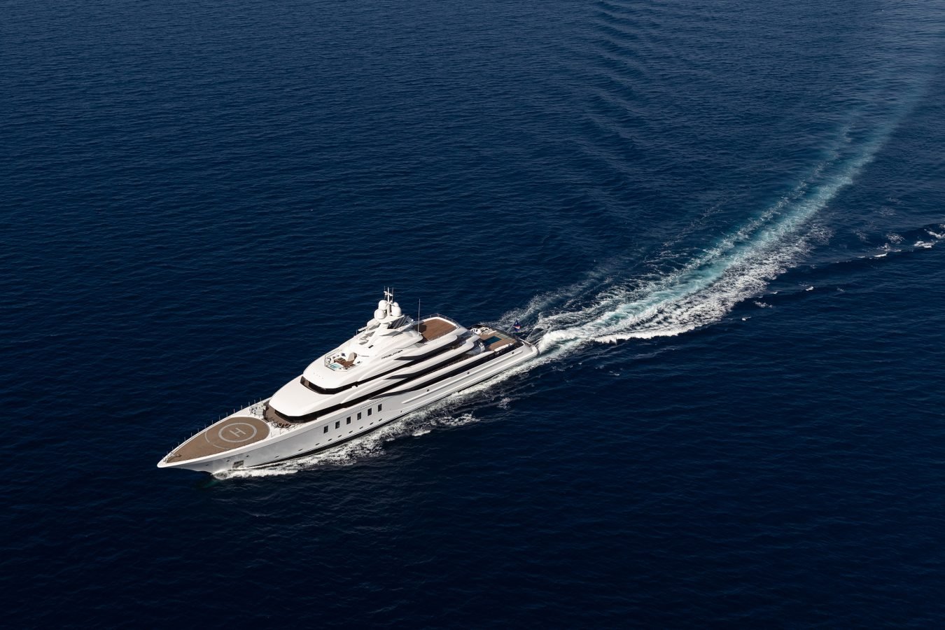 A large white luxury yacht with a helipad sails through deep blue ocean, leaving a wake behind it.