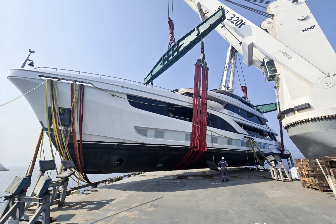 A white Majesty 145 superyacht suspended by heavy red straps from a crane, with a lone worker standing on the deck below.