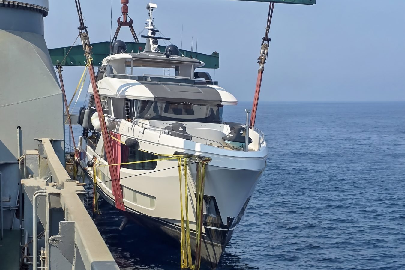 A Majesty 145 superyacht is suspended by crane straps above the blue ocean, framed by a large metal structure.
