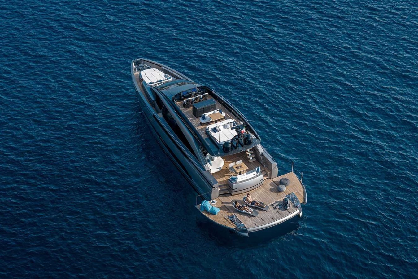 The Pershing Gtx80 luxury yacht floats on calm blue water; several people relax on its deck under sunny skies.