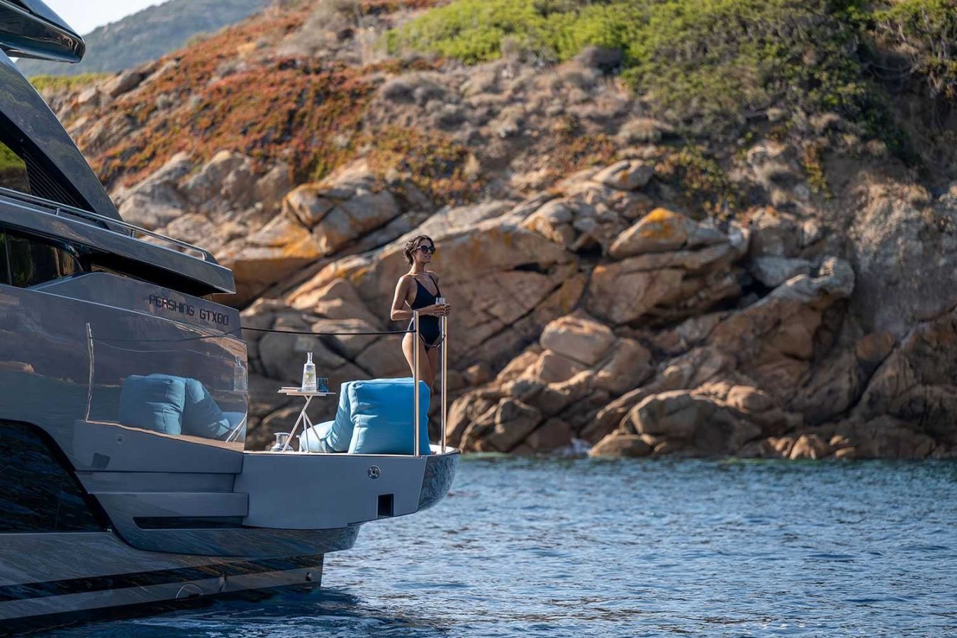 Woman in a black swimsuit stands on Vicky yacht’s deck, with rocky shoreline and greenery in the background.