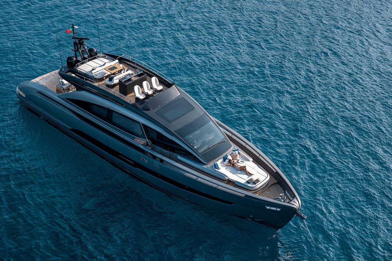 A sleek Pershing Gtx80 yacht with lounge areas floats on deep blue water under clear skies, viewed from above.