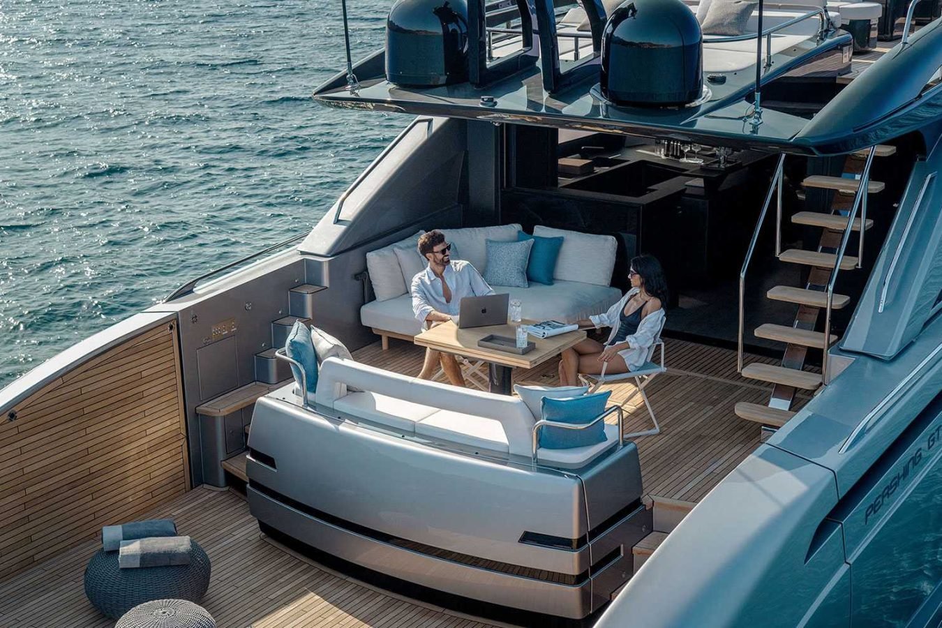 Two people work on a laptop aboard a Pershing Gtx80 yacht for charter, enjoying blue waters and luxe deck seating.