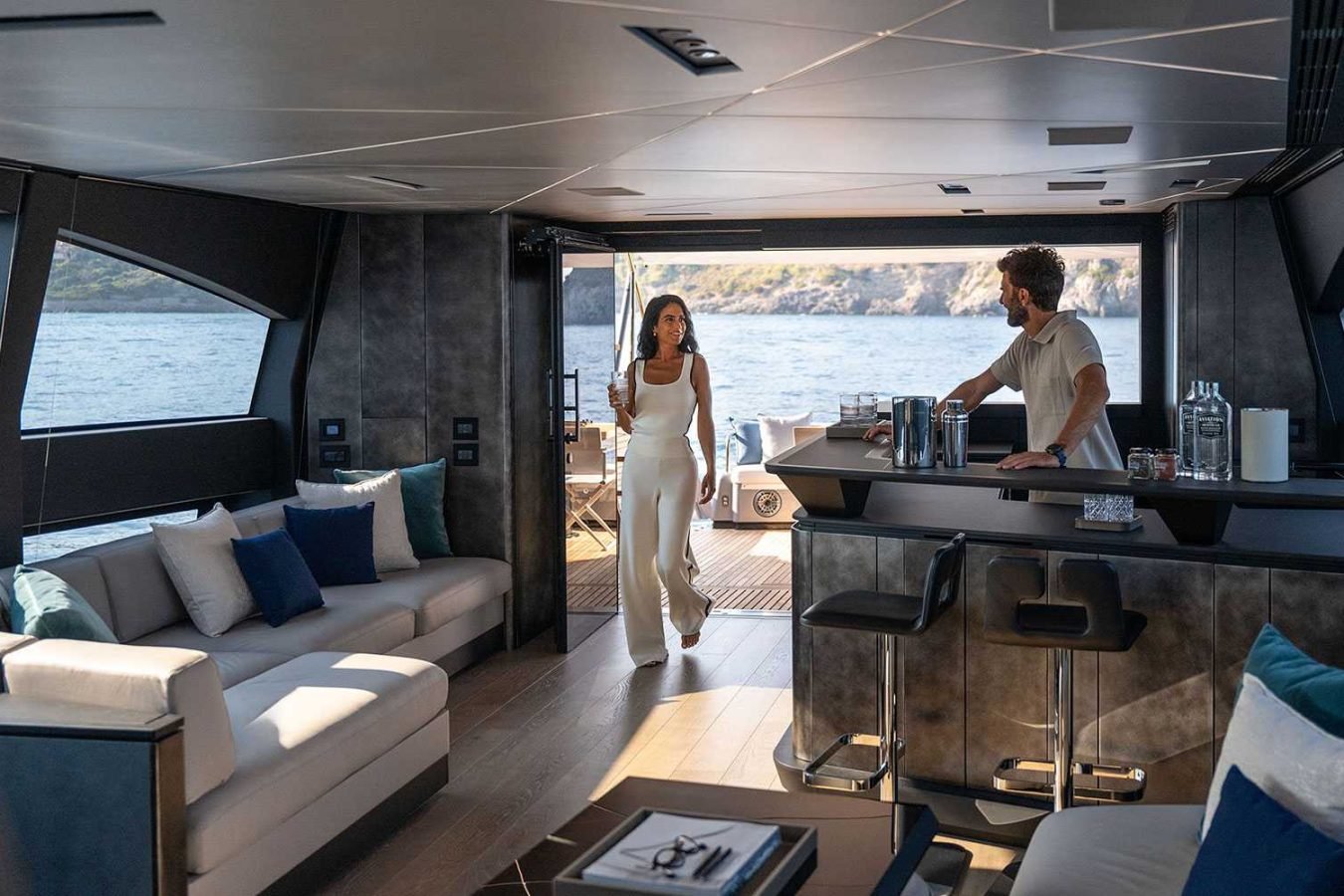 A woman in white walks toward a man at a modern Pershing Gtx80 yacht bar; ocean and cliffs visible through open doors.