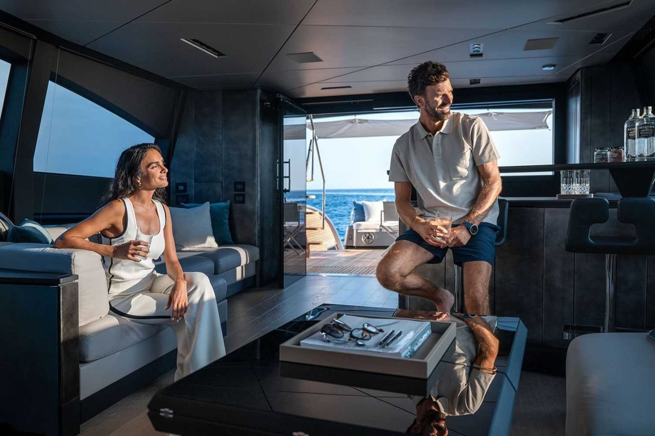 A smiling couple relaxes with drinks on a Pershing Gtx80 yacht for charter, enjoying the modern lounge and ocean views.