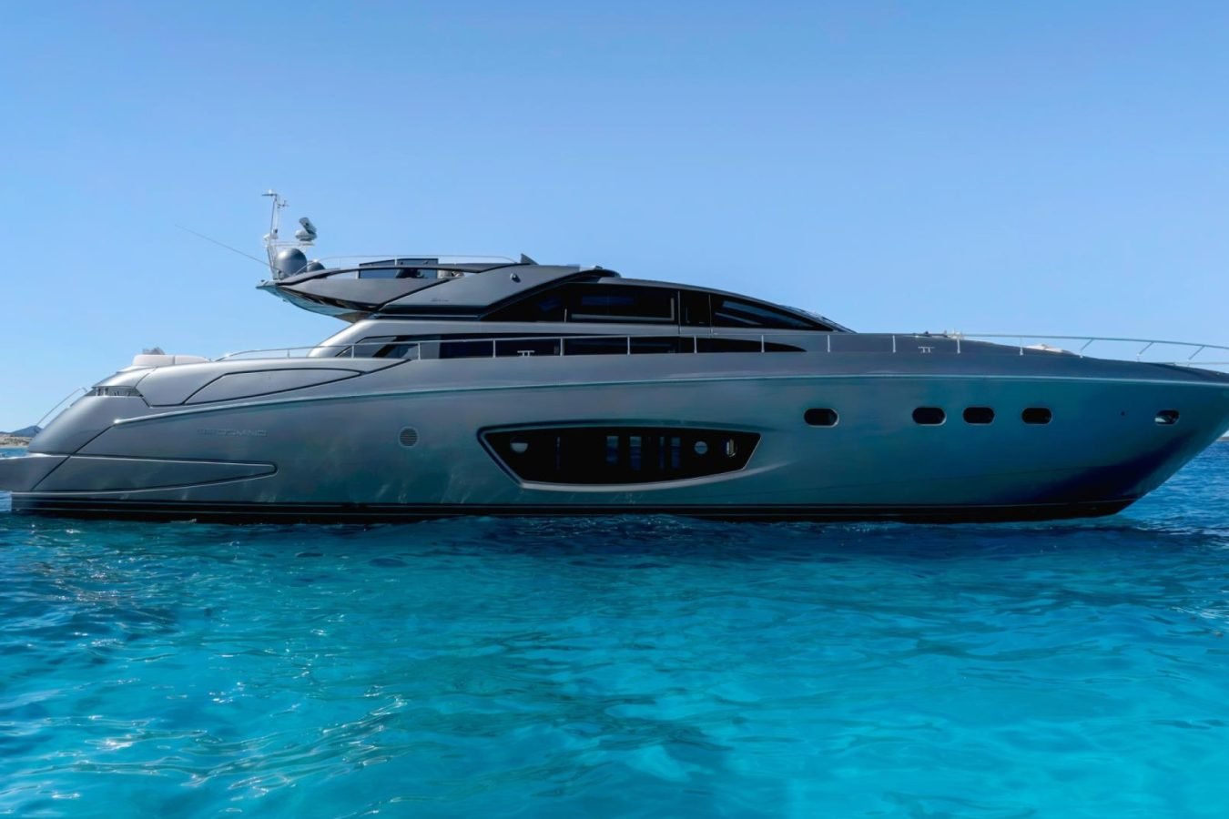 Sleek, modern Riva 86 Tanit yacht floats on clear blue water under a bright blue sky, viewed from the side.