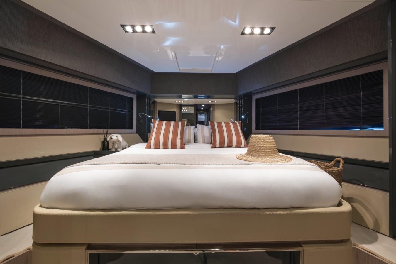 A Riva 86 Tanit yacht bedroom with a large bed, striped pillows, a straw hat, soft lighting, and dark window blinds.