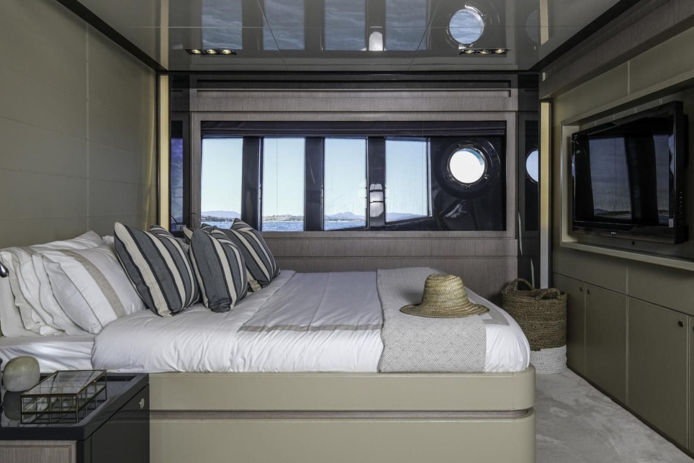 Modern Riva 86 yacht bedroom with striped pillows, straw hat on bed, large windows, and wall-mounted TV.