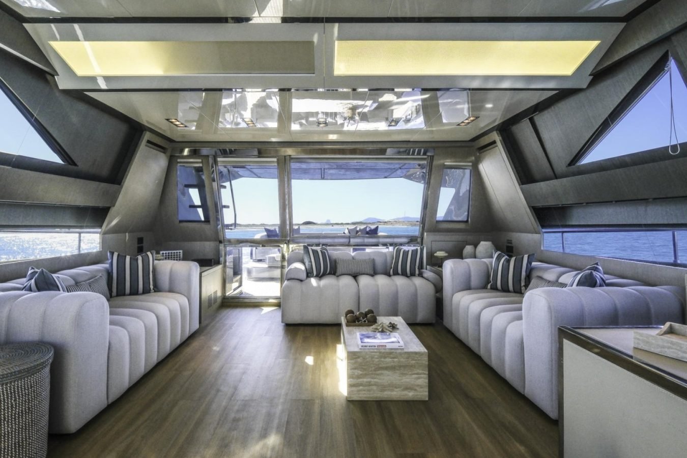 Modern yacht for charter with plush gray sofas, striped pillows, low table, large windows, and stunning ocean view.