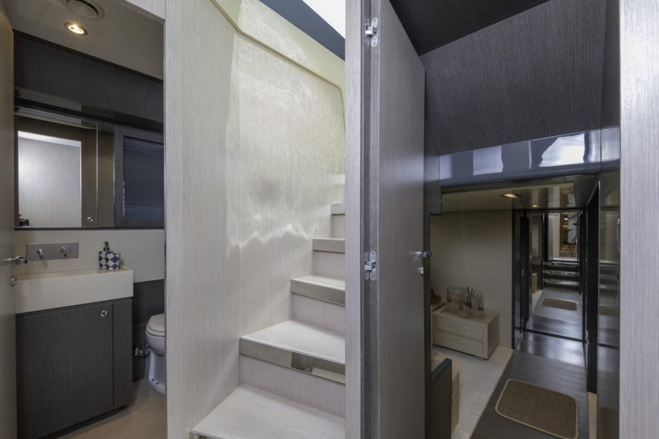 Modern Riva 86 yacht for charter; light wood stairs, doors to bathroom left, hallway with rug and Tanit seating area right.