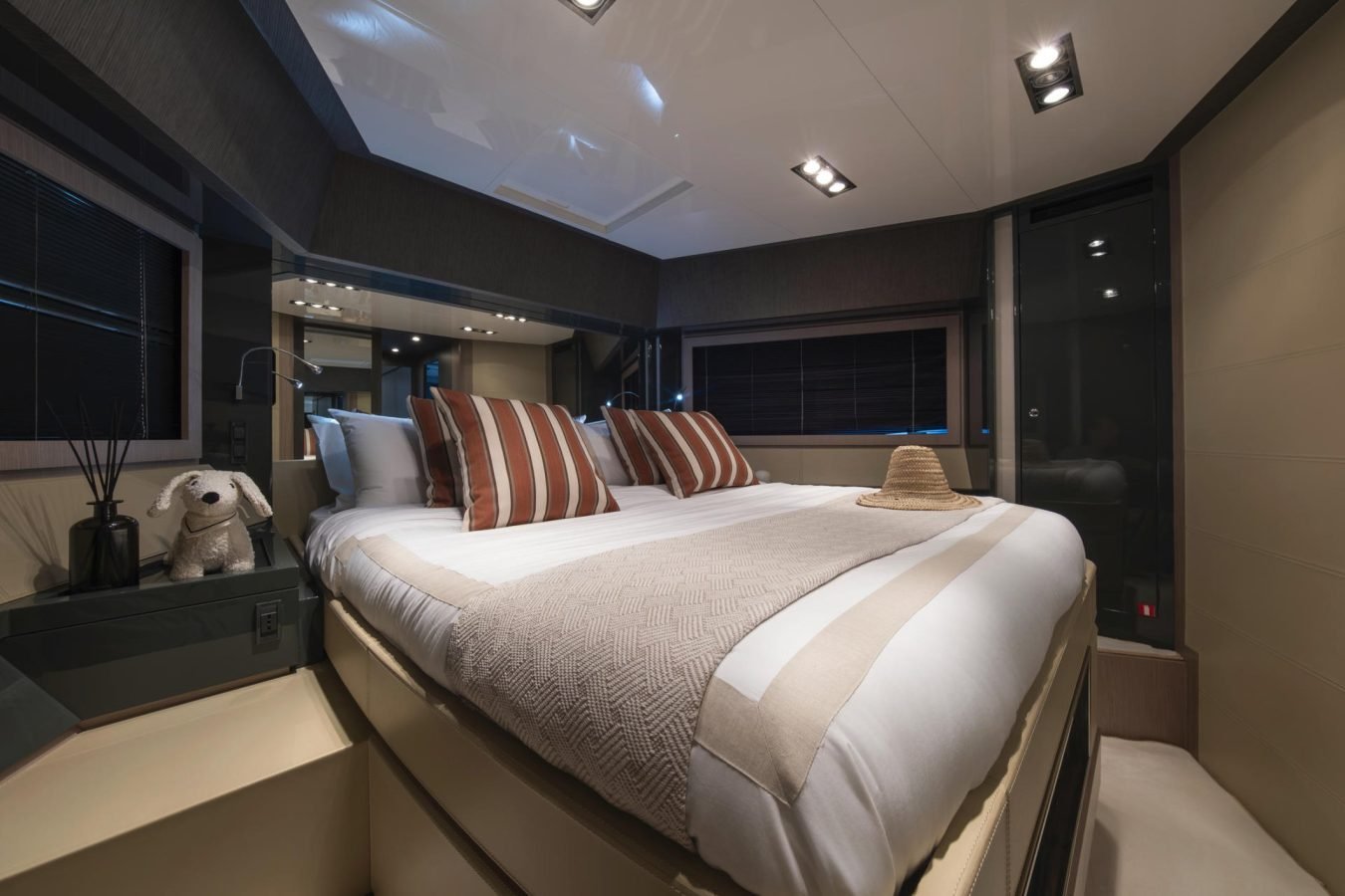 Modern bedroom on a Riva 86 Tanit yacht for charter, featuring striped pillows, a beige throw, and a stuffed dog.