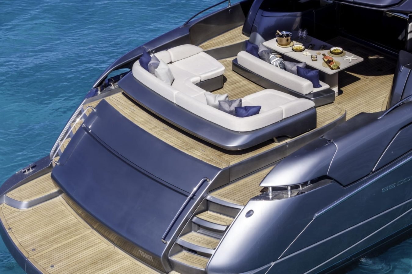 Luxury Riva 86 Tanit yacht for charter features wood deck, sun loungers, dining table, and blue pillows on clear water.