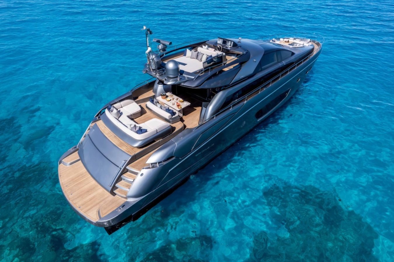 A Riva 86 Tanit luxury yacht floats on clear blue water, revealing wooden decks, lounge areas, and ocean views below.