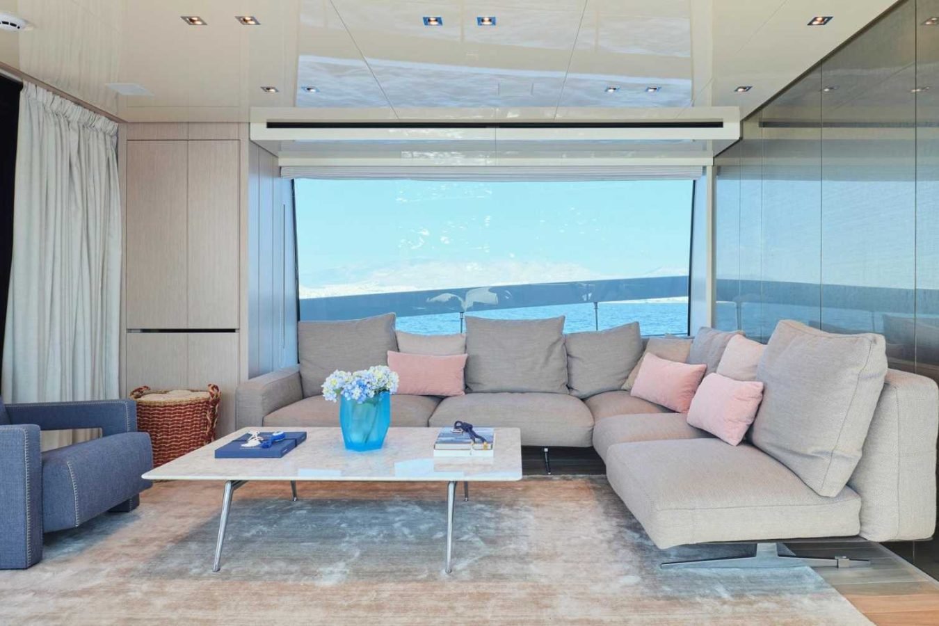 Modern yacht lounge in Sanlorenzo SX76 Fuquet Duet with beige sectional, pastel pillows, blue armchair, and ocean view.