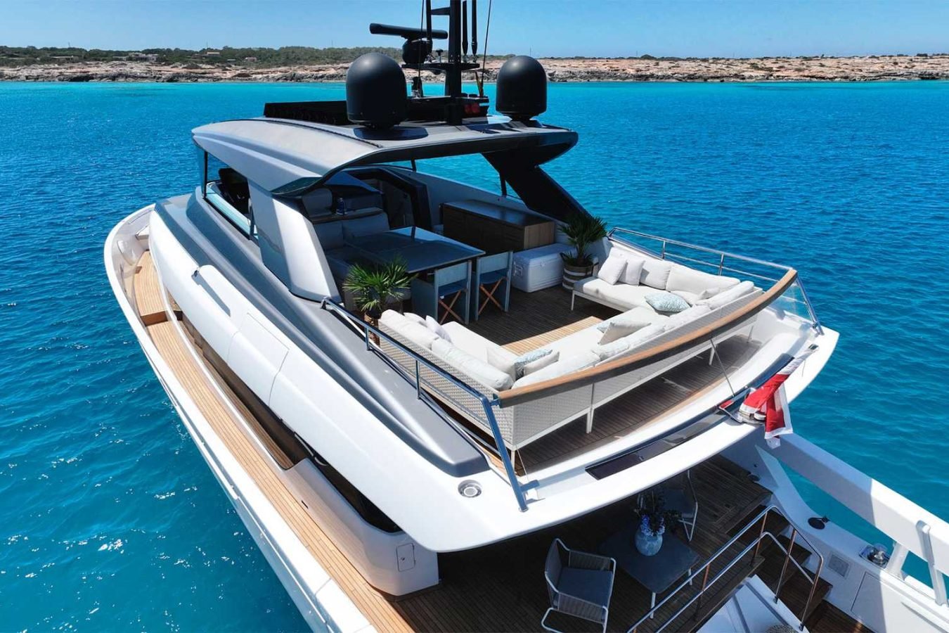 The Sanlorenzo SX76 Fuquet Duet yacht, with sun loungers and potted plants, sails near a rocky shoreline.