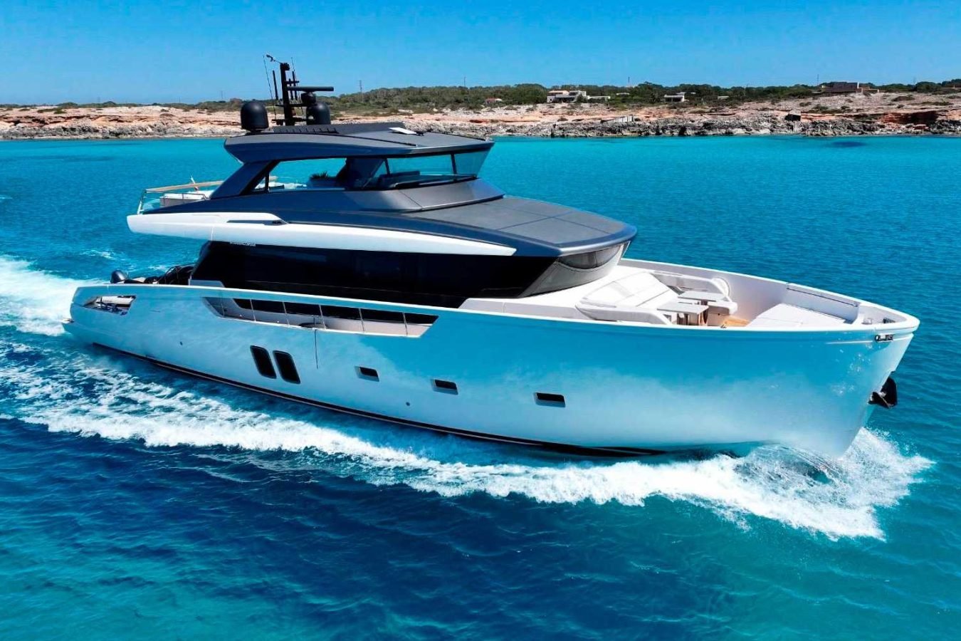 The Sanlorenzo SX76 Fuquet Duet, a sleek white yacht, sails on clear blue water near a rocky coastline under a cloudless sky.