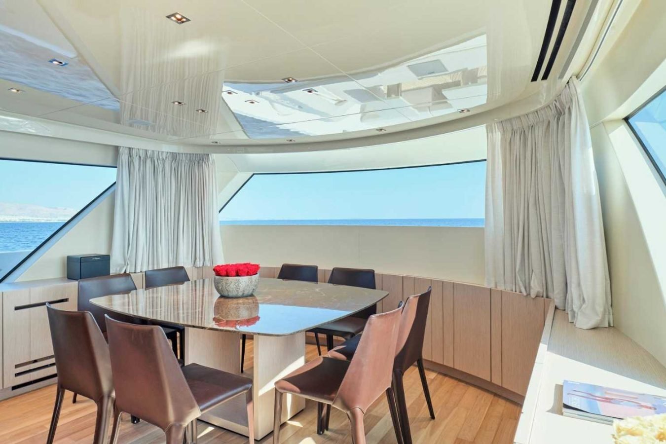 Modern Sanlorenzo SX76 Fuquet Duet yacht dining area with large windows, round table, and red roses in sunlight.