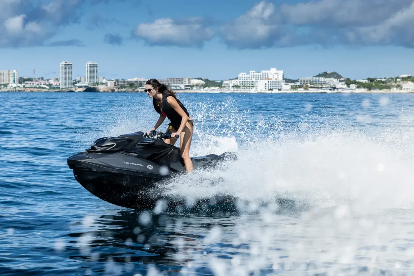 Woman in swimsuit rides black jet ski on blue water near city, with Lurssen CC-Summer yacht for charter in the background.