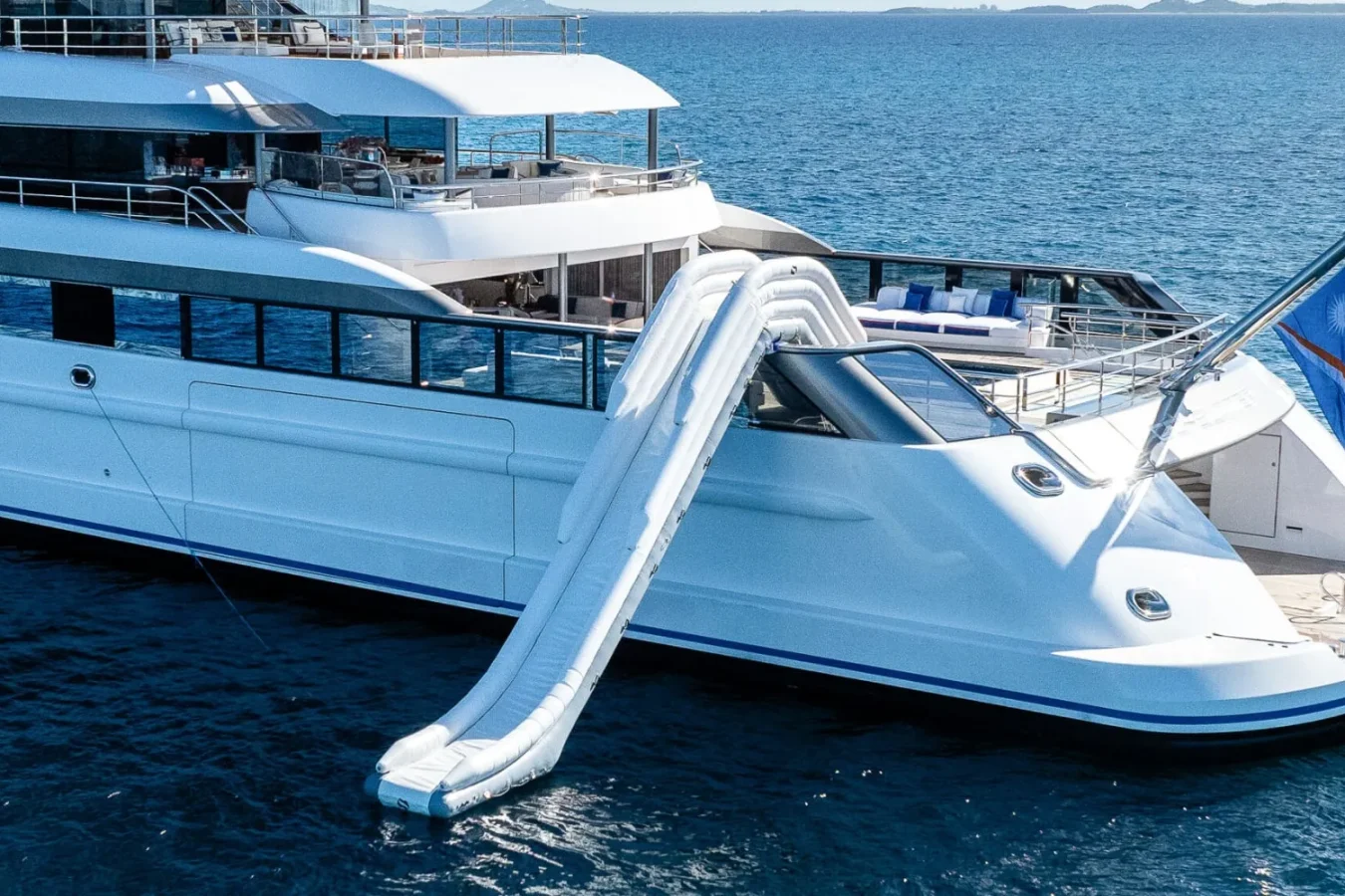 Luxury Lurssen CC-Summer Yacht for Charter with an inflatable slide from the upper deck into the blue ocean, flag at stern.