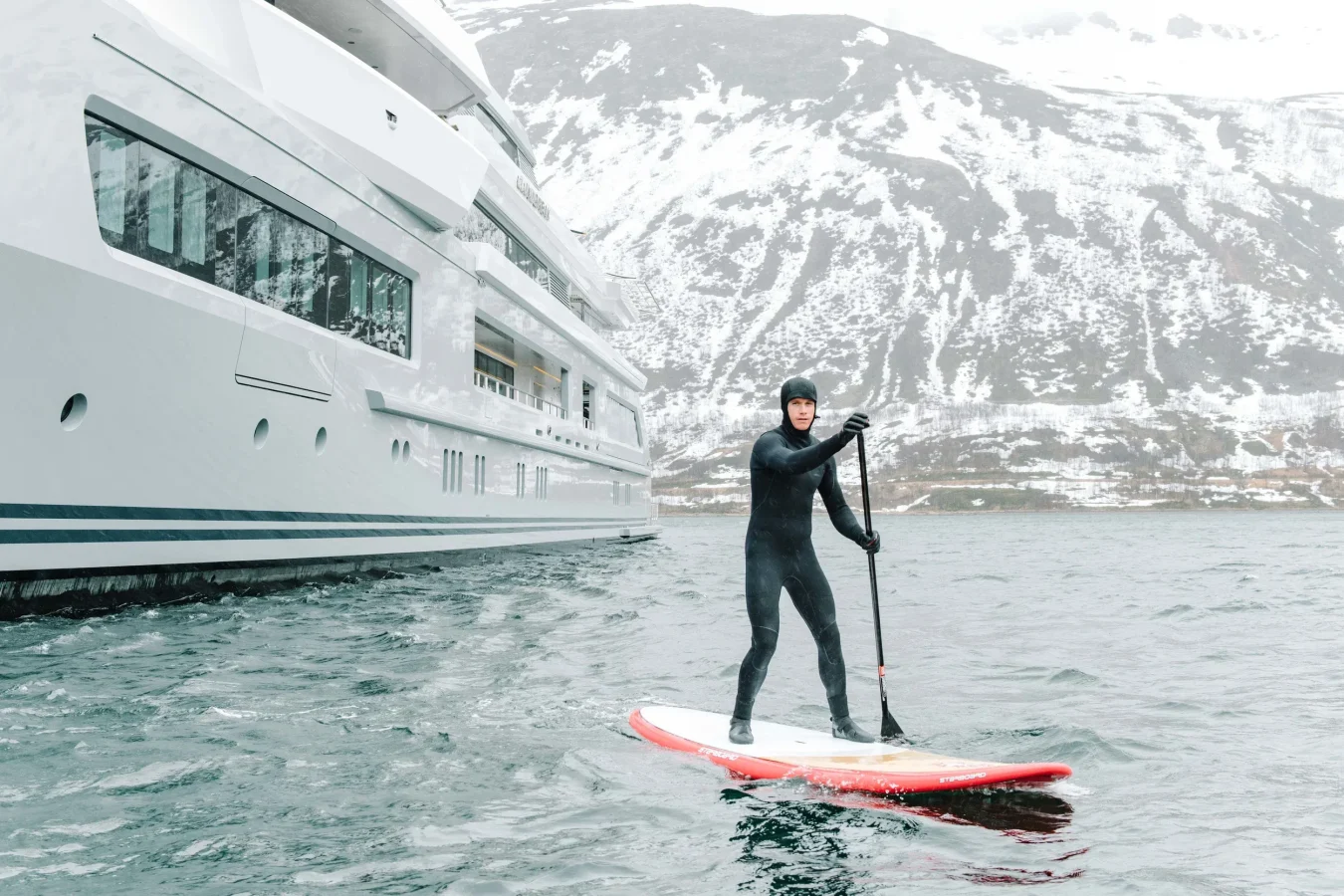 A person in a wetsuit paddleboards near the Abeking Rasmussen Cloudbreak yacht for charter with snowy peaks behind.