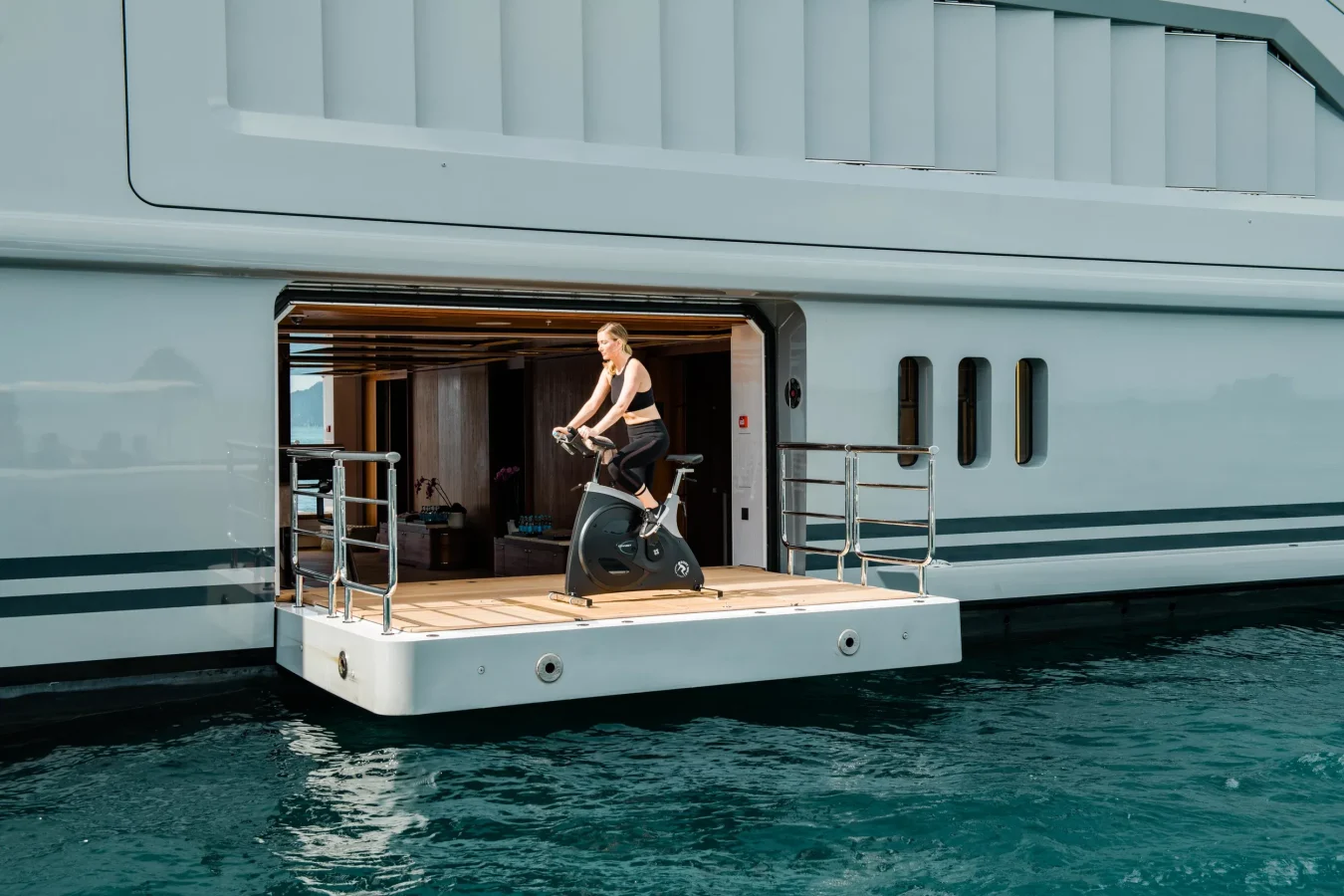 A person rides an exercise bike on the Abeking Rasmussen Cloudbreak yacht for charter, overlooking calm ocean water.