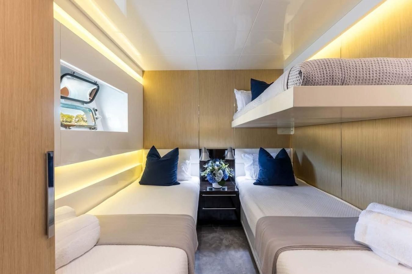 Modern yacht cabin aboard Arcadia 85 with twin beds, upper bunk, blue pillows, flowers, window, and soft lighting.