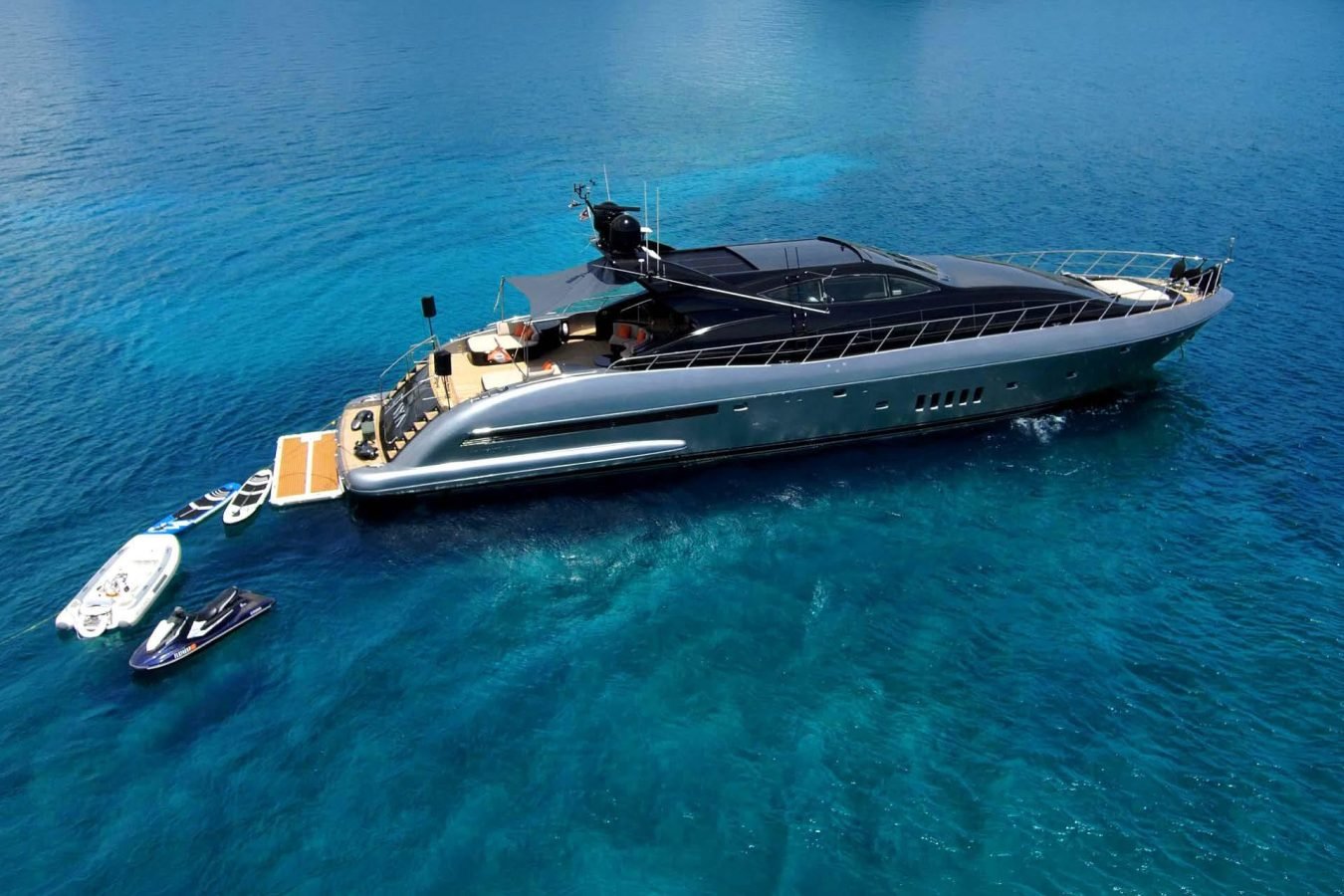 The Mangusta 105 Daya Yacht is anchored in clear blue water, with jet skis and a small boat nearby on the right.