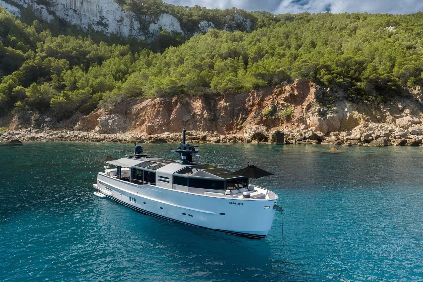 The Arcadia 85 Dhamma II yacht for charter floats on clear blue water near a rocky, tree-covered coastal cliff.