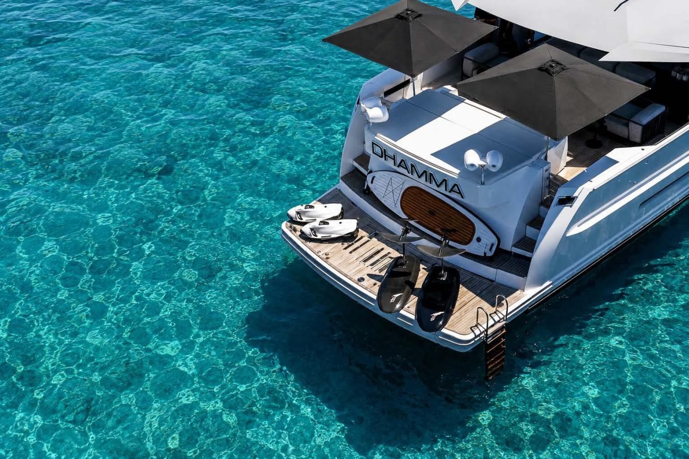 A luxury Arcadia 85 yacht, DHAMMA, anchored in blue water, features water toys and two black umbrellas on the deck.