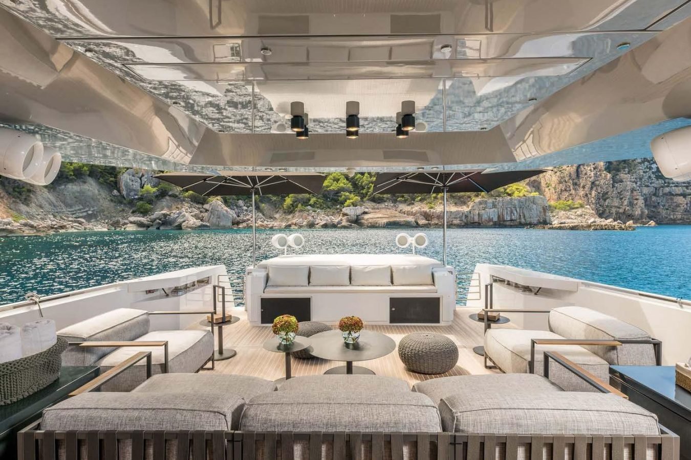 Luxurious Arcadia 85 yacht deck with modern seating, tables, and ocean view; rocky shoreline and greenery in background.
