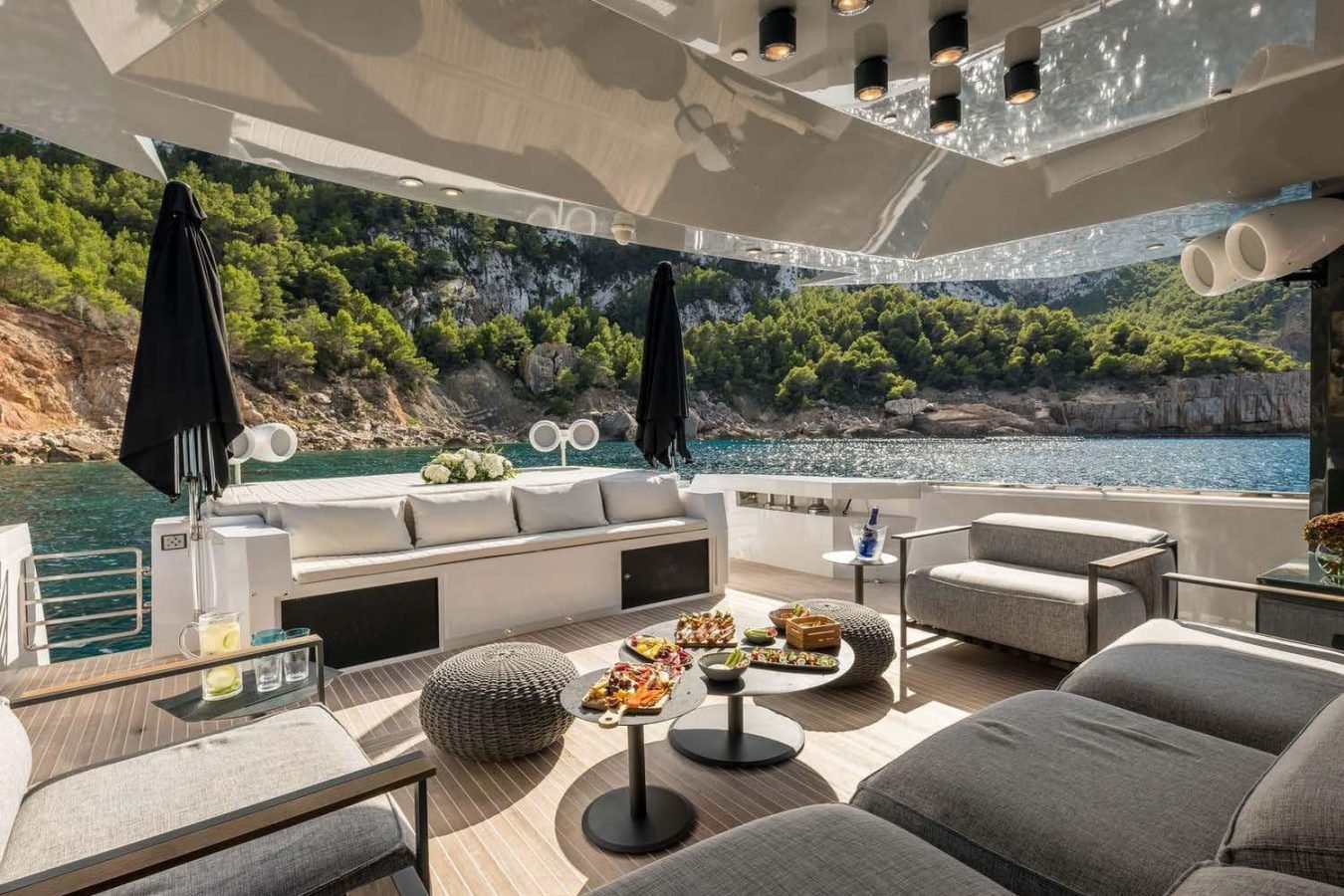 Luxury Arcadia 85 yacht deck with gray sofas, snacks, black umbrellas, and scenic rocky shoreline in the background.