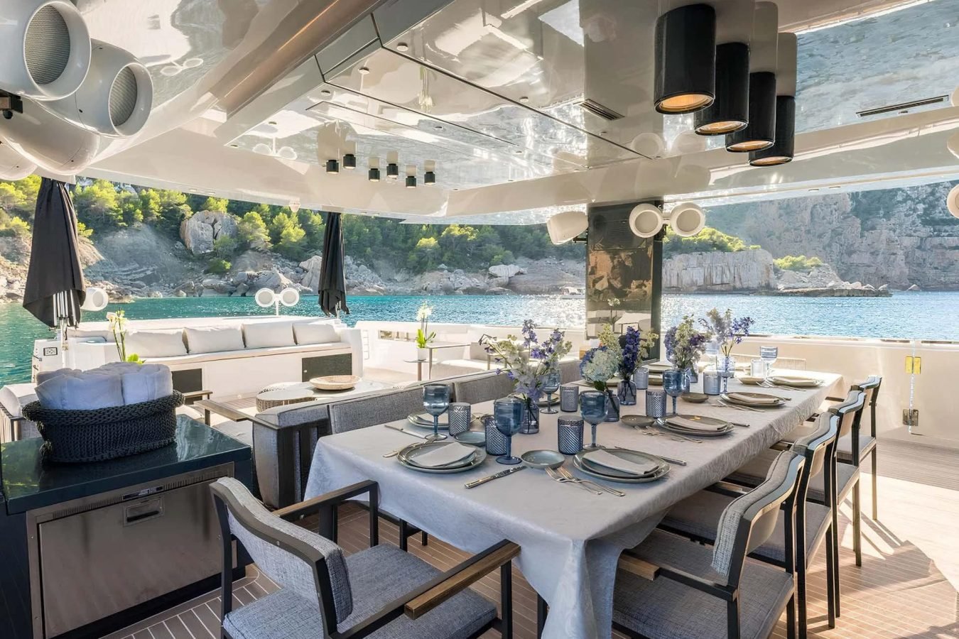 Elegant Arcadia 85 yacht dining area set for a meal with blue accents, overlooking clear water and rocky, tree-lined shore.
