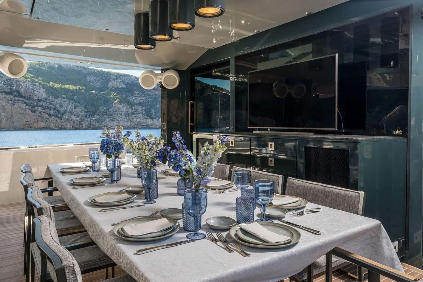 Elegant Arcadia 85 yacht dining area set for eight, with blue glassware, florals, and a scenic sea view.
