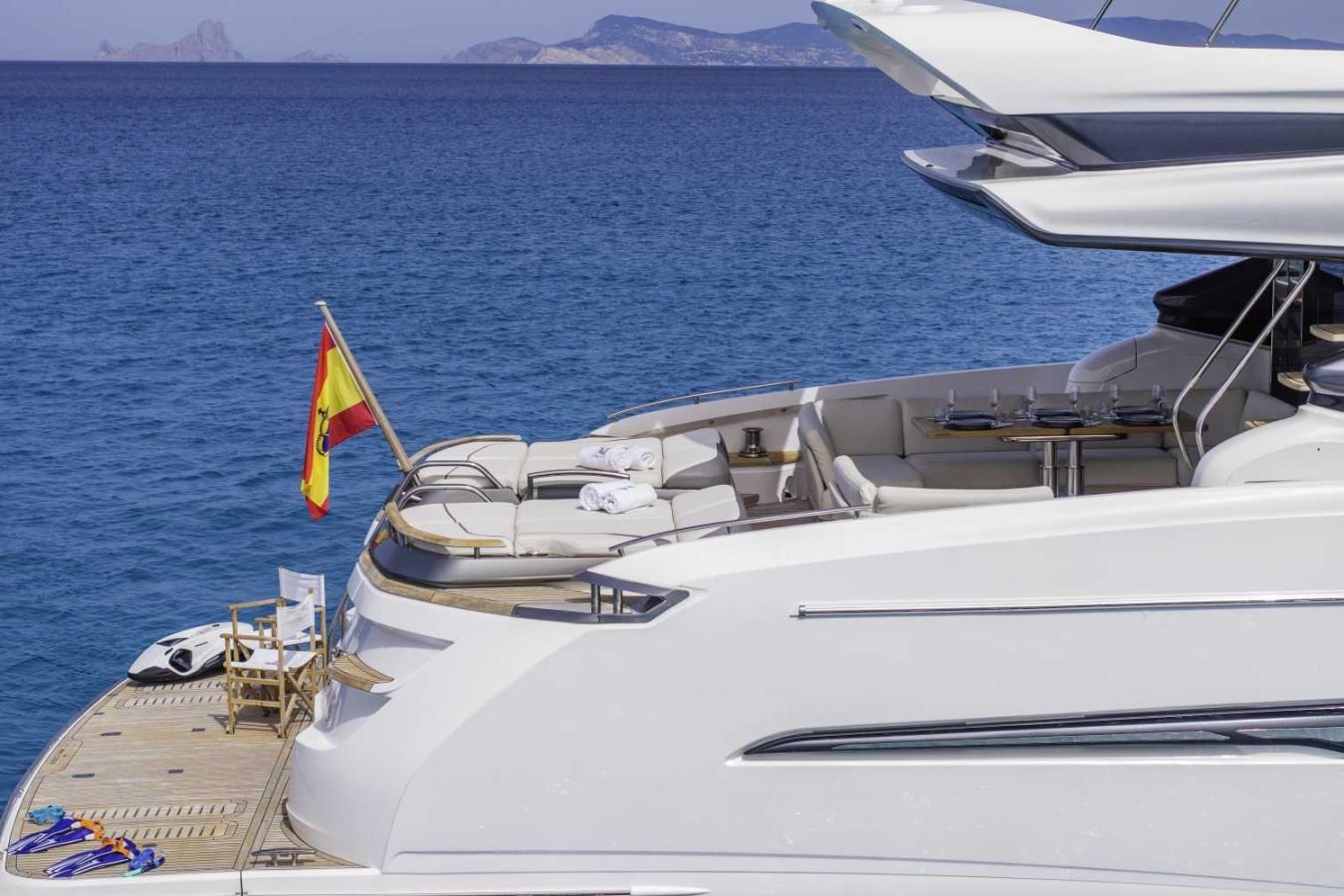 A white Princess S78 yacht with a Spanish flag floats on blue water; mountains and sky form the distant background.