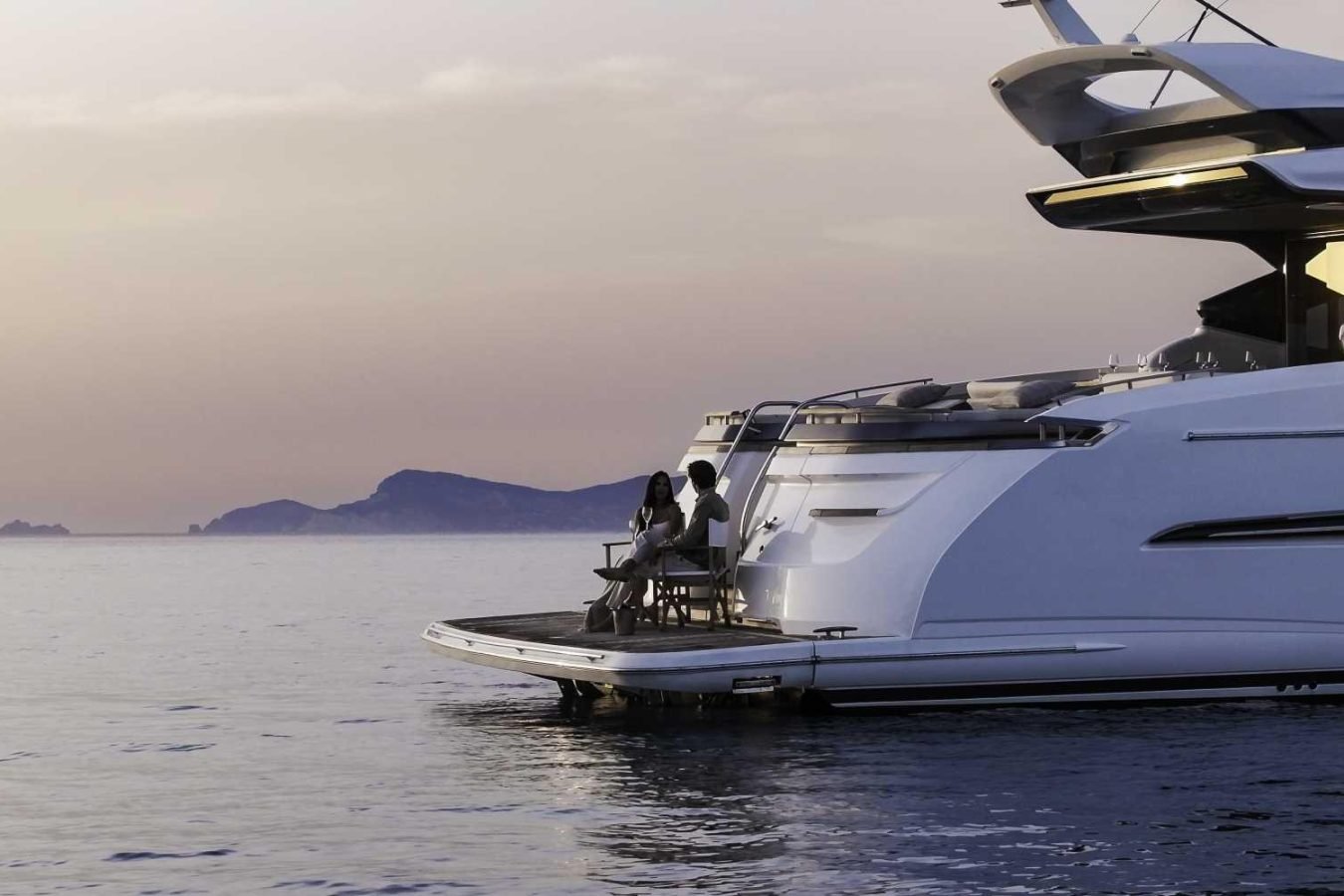 Two people relax on the deck of Why Knot, a Princess S78, as the sun sets over calm water and distant islands.