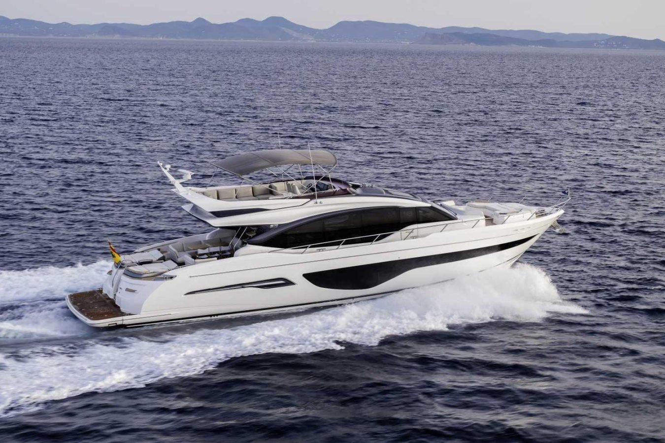 The Princess S78 Yacht for Charter glides swiftly through blue water, leaving a white wake; hills line the horizon.