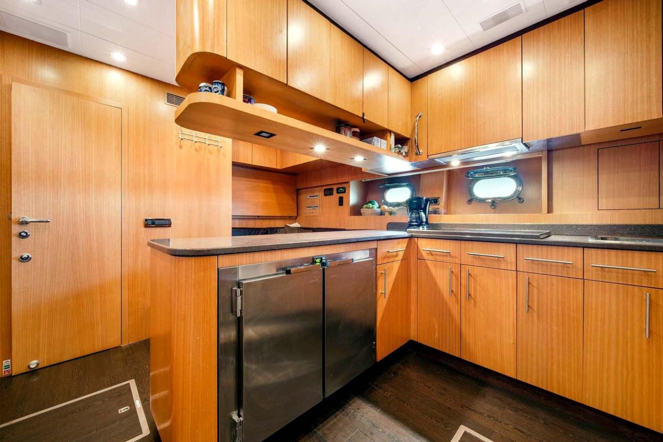 Modern Mangusta 105 yacht kitchen with wood cabinets, black counters, stainless fridge, and porthole windows above sink.