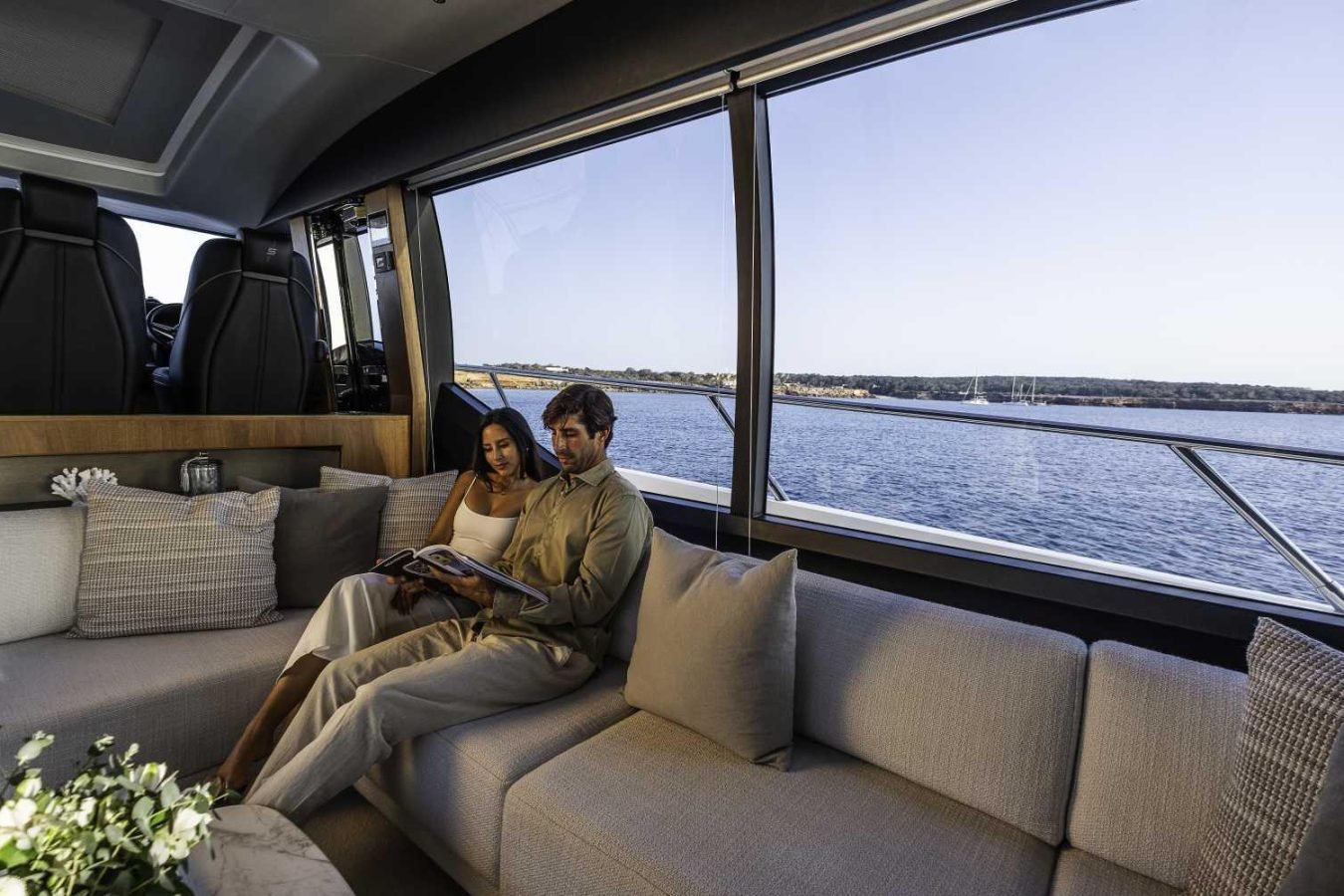 A couple sits on a Princess S78 yacht’s sofa reading, with calm water and a distant shore visible through large windows.
