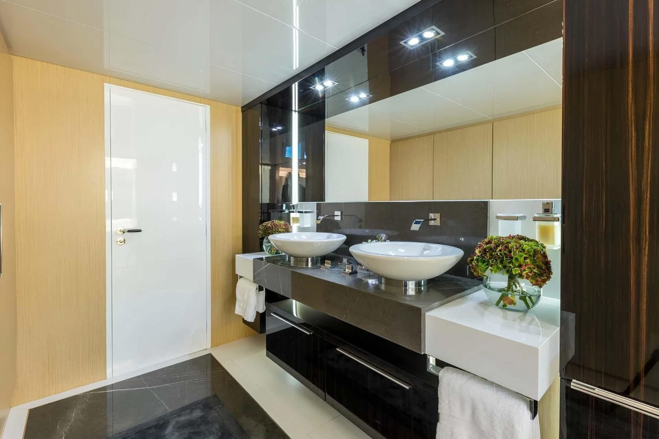 Modern bathroom on the Dhamma II yacht for charter, with vessel sinks, dark vanity, mirror, flowers, and beige walls.