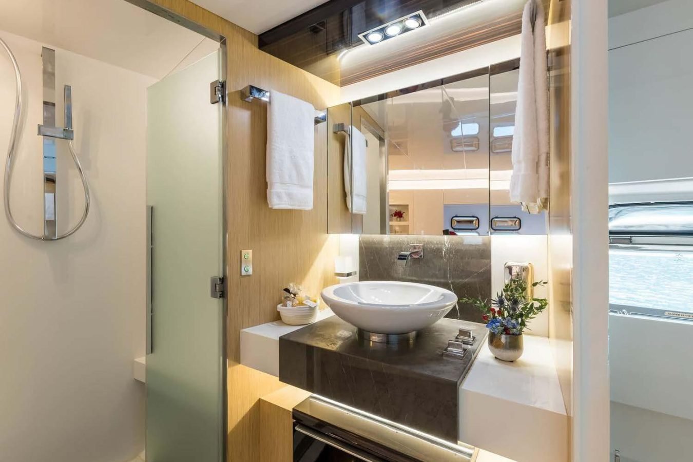 Modern Dhamma II yacht bathroom with vessel sink, mirror, towels, glass shower door, and window showing water outside.
